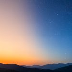 A wide sky transitioning from a warm orange sunrise on the left to a deep blue starry night on the right over silhouetted hills