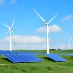 Blue solar panels and tall white wind turbines on green rolling hills under a bright blue sky